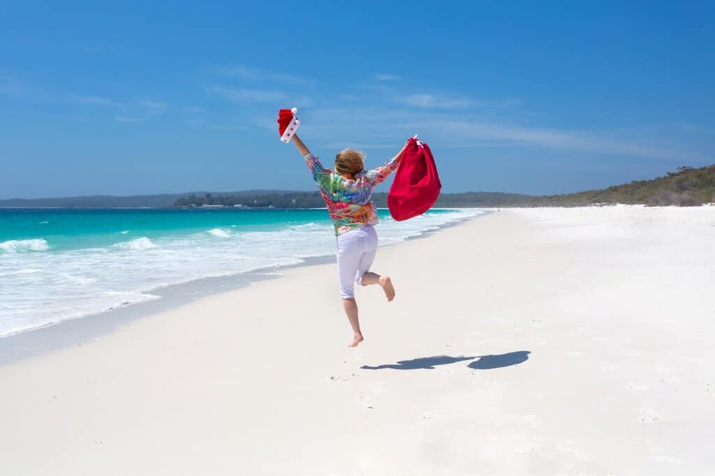 A person running along a beach holding a Santa hat and red bag, celebrating Christmas under the summer sun. One of the fun Australian Christmas facts is that many people celebrate the holiday outdoors, making the most of the warm weather and stunning beaches.