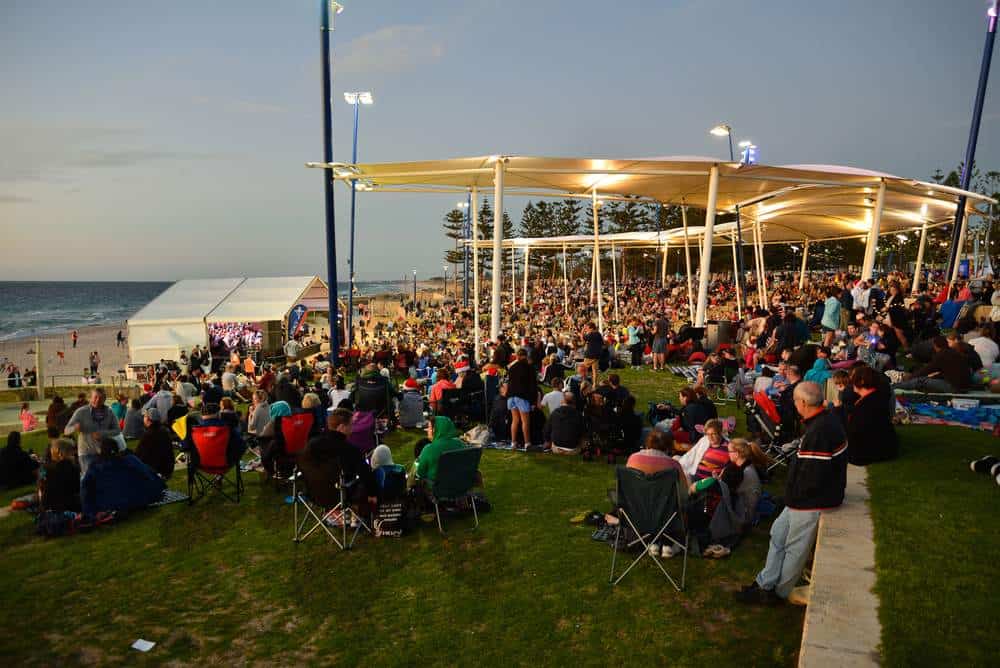 Large crowd gathered by the beach at an outdoor Christmas carol event in Perth, with families enjoying festive music and holiday celebrations under a lit pavilion. Highlights the community spirit and family-friendly atmosphere of Christmas in Perth.