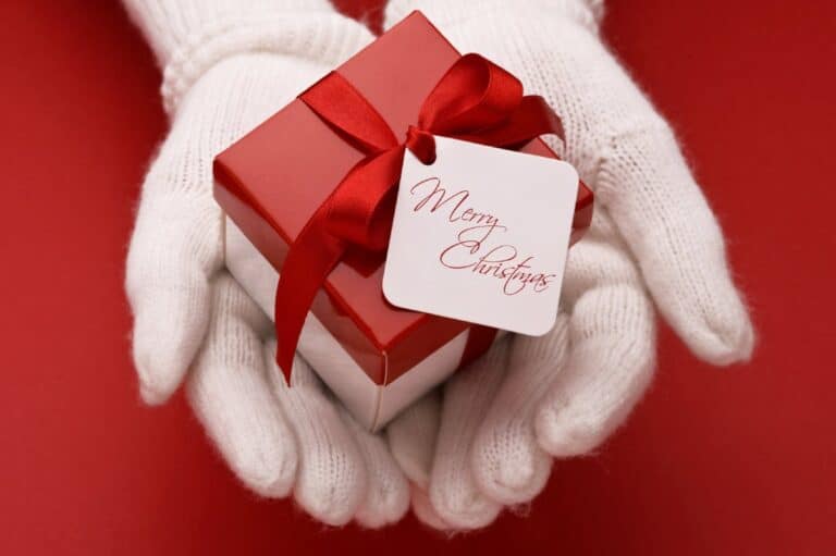 Hands in white gloves holding a red Christmas gift box with a red ribbon and a tag that says 'Merry Christmas.' This image relates to the blog post about finding the perfect Christmas present for mum, showcasing a beautifully wrapped gift symbolising thoughtful holiday gift-giving.