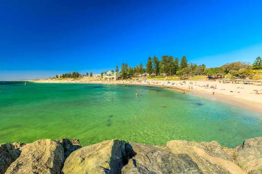Clear turquoise waters and sunbathers at Cottesloe Beach, a family-friendly destination among the beaches in Western Australia, with the iconic Indiana Tea House in the background.