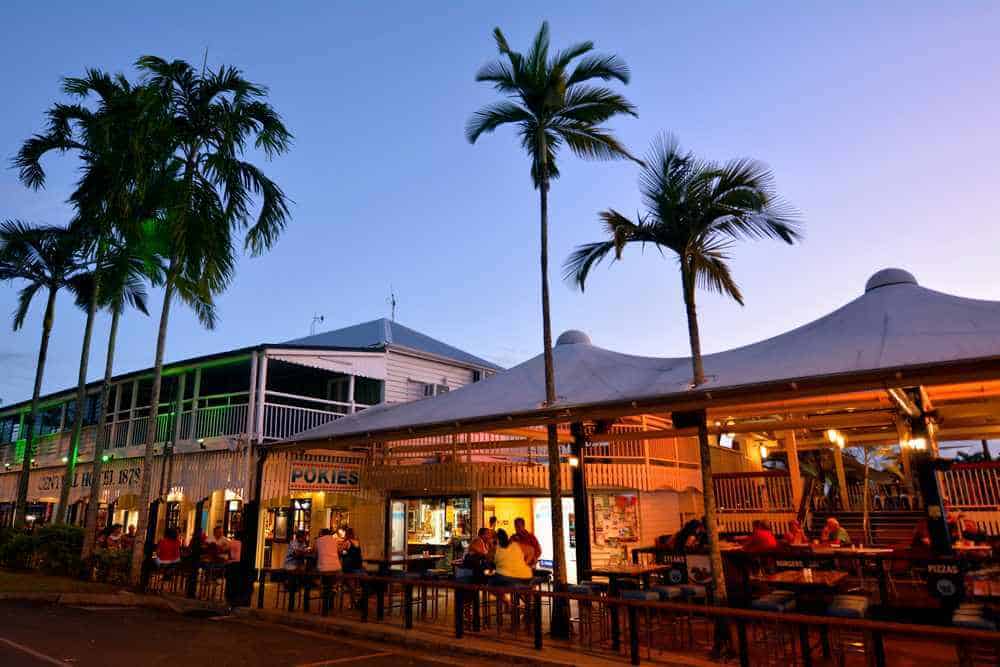 Evening falls on a lively outdoor dining scene in Port Douglas, with people relaxing at tables under warm lighting, palm trees swaying above. This social hotspot exemplifies the inviting atmosphere of Port Douglas, contrasting with the serene, natural beauty of the Whitsundays, both jewels in the crown of Queensland's holiday destinations