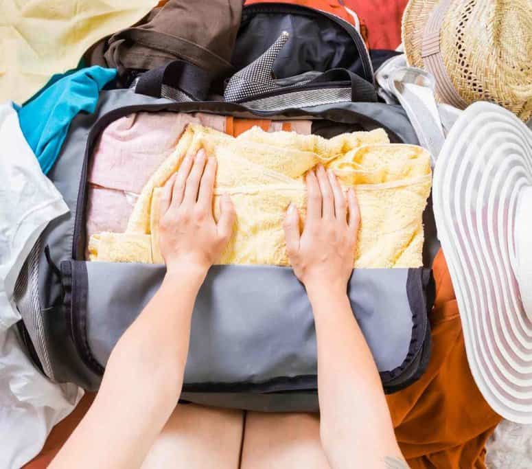A person packs a gray suitcase with neatly folded clothes, including a yellow towel. Their hands press down on the clothes. Surrounding the suitcase are a hat with a wide brim and a woven straw hat.