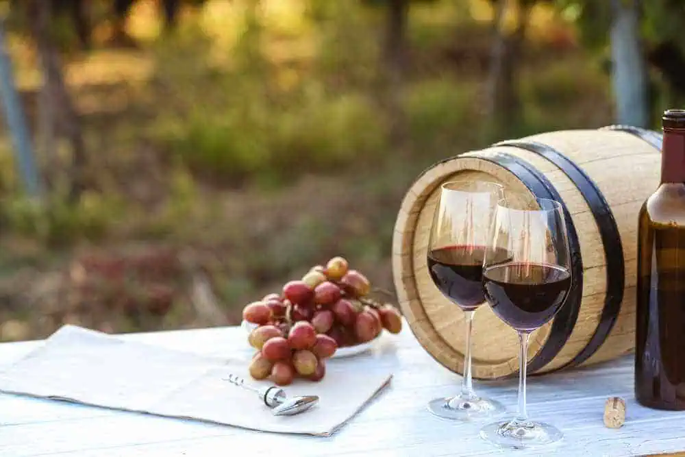 Two glasses of red wine set before an oak barrel, with a bunch of grapes and a wine bottle, a serene scene typical of a wine tasting tour in Hunter Valley.