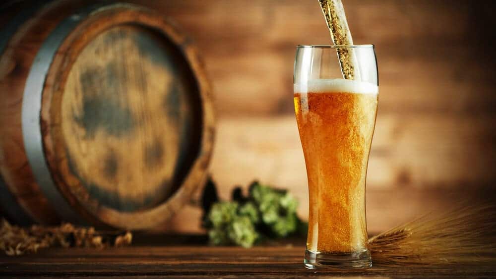 A tall glass of frothy beer being poured against a rustic backdrop with a wooden barrel and hops, illustrating the traditional brewing methods explored on brewery tours in Brisbane.