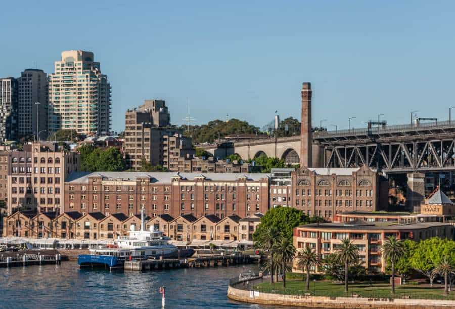 View of The Rocks district in Sydney, with historic warehouses converted into modern establishments along the waterfront, and the Sydney Harbour Bridge in the background. The area’s rich history is often highlighted in chilling tales on Sydney ghost tours.