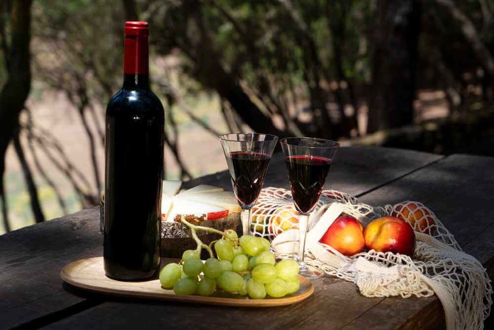 A bottle of wine with two glasses, cheese, grapes, and peaches on a table, set against a tranquil forest backdrop, reminiscent of the best Hunter Valley wine tours.