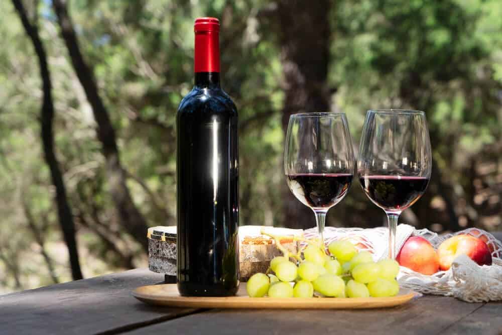 A bottle of red wine with two filled glasses, accompanied by grapes and cheese on a wooden table, set against a natural forest backdrop, ideal for a Hobart wine tasting excursion.