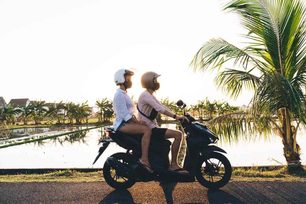 Two people riding a hired scooter at sunset near a palm-fringed rice field in Bali, a popular alternative to Uber for exploring the island's beauty at one's own pace.