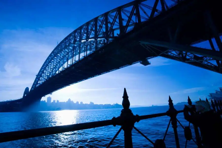 Silhouette of Sydney Harbour Bridge against a bright blue sky, with the sun reflecting off the water, creating a serene yet mysterious atmosphere. This iconic structure is not only a landmark but also a fixture in the stories shared on Sydney ghost tours.