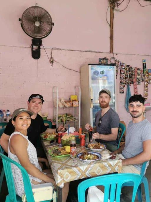 Four friends enjoying a meal at Pink Warung, a local eatery in the Gili Islands, with a casual setting and a wall adorned with colourful postcards. Sharing a meal here is a relaxing and authentic thing to do in the Gili Islands, perfect for travellers seeking cultural experiences