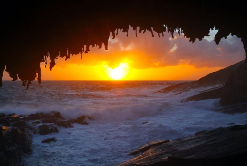 Golden sunset viewed from a cave in Flinders Chase National Park, Kangaroo Island, with waves crashing against the rocky shore. This image highlights one of the best places to watch sunsets in Australia, supporting the blog post about must-see sunset destinations.