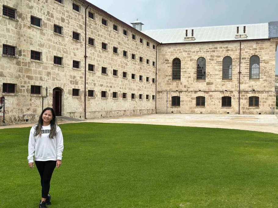 Woman standing on the green lawn of Fremantle Prison in Australia, smiling at the camera. The historic building, known for its limestone walls and barred windows, is in the background. This image illustrates the unique attractions in Australia, helping readers answer the question "Is Australia worth visiting?" by showcasing its rich historical sites.