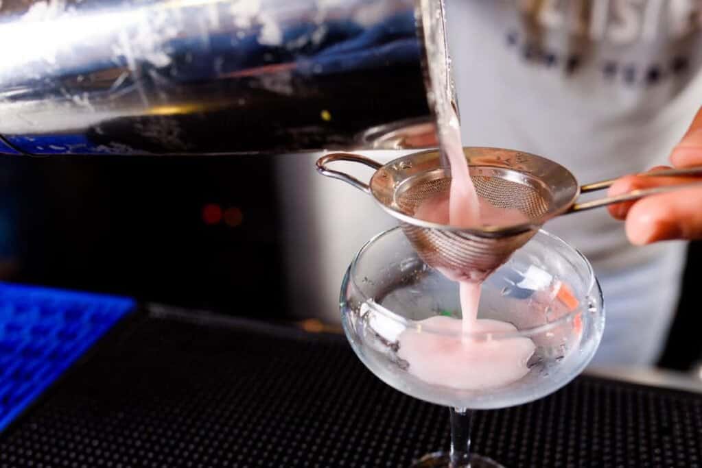 A bartender pouring a pink gin cocktail through a strainer into a glass. This image represents a mixology experience.