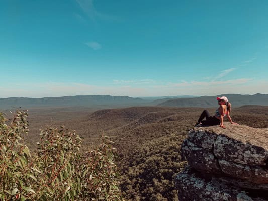 A hiker bends down to adjust their shoe on a prominent rocky outcrop along the Grampians Peaks Trail, overlooking the expansive forest-covered hills under a clear blue sky.