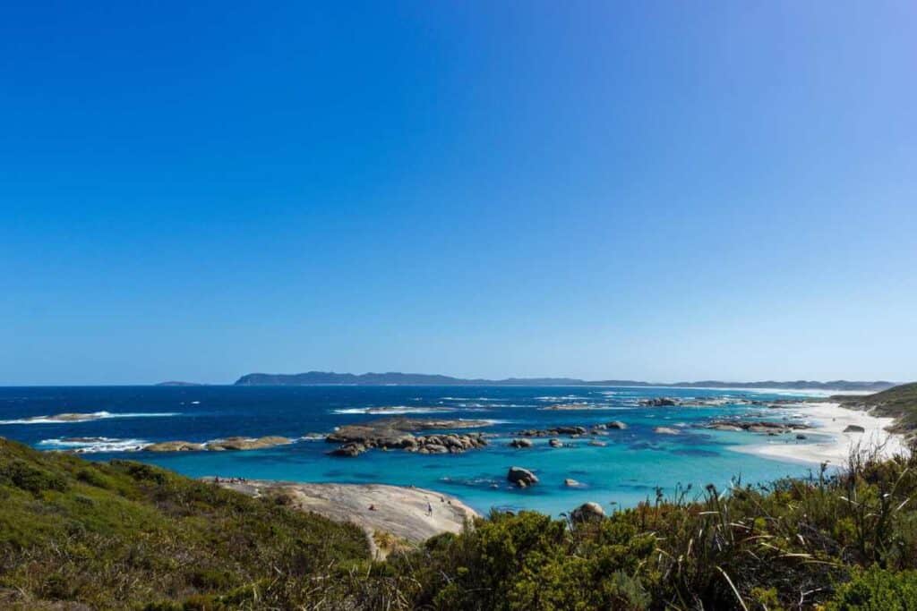 A panoramic view of Greens Pool, heralded as one of the most beautiful beaches in Western Australia, showcasing clear turquoise waters, scattered rock formations, and white sandy beaches framed by lush greenery.