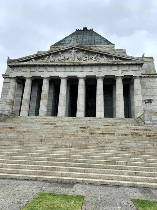 A large stone building with a pyramid-shaped roof, featuring six tall columns at the entrance and a set of wide steps leading up to it, should be part of any Melbourne itinerary. The structure has detailed carvings above the columns, set against an overcast sky.