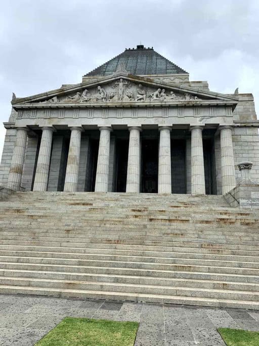 A large stone building with a pyramid-shaped roof, featuring six tall columns at the entrance and a set of wide steps leading up to it, should be part of any Melbourne itinerary. The structure has detailed carvings above the columns, set against an overcast sky.