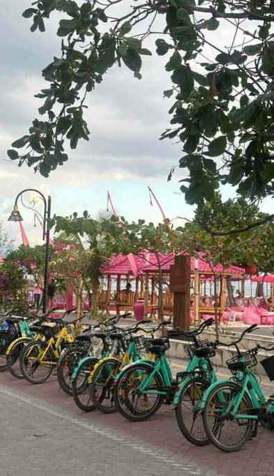 Row of colorful bicycles lined up for rent on a promenade in the Gili Islands, with a backdrop of a vibrant beach cabana with pink cushions. Biking is a popular and eco-friendly activity among the many things to do in Gili Islands, perfect for exploring the scenic beauty of the area.