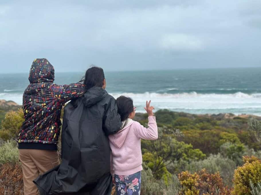 Our kids in warm jackets standing on the sand dunes near Waitpinga beach campground, facing the wild Southern Ocean and waving at the waves—capturing a candid moment from a windswept family camping trip on the Fleurieu Peninsula.