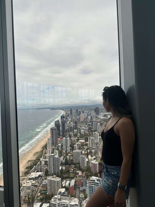 The author, Kate, standing inside a high-rise building gazes out at the sweeping cityscape of Gold Coast in December, dressed in summer casuals suitable for the warm climate.