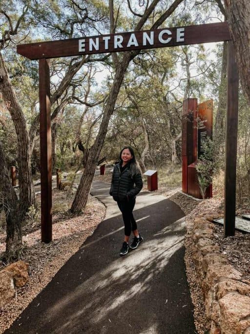 A woman stands at the entrance of Ngilgi Cave, surrounded by trees and natural scenery. She is smiling and wearing a black jacket and black leggings. This image highlights the inviting entrance to Ngilgi Cave, supporting the post: Is Ngilgi Cave worth visiting?