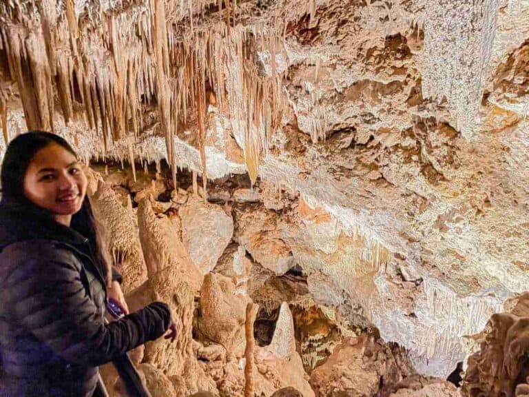 A woman smiles while standing inside Ngilgi Cave, with intricate stalactites and stalagmites visible in the background. This image supports a detailed Ngilgi Cave review by showcasing the cave's stunning interior formations.