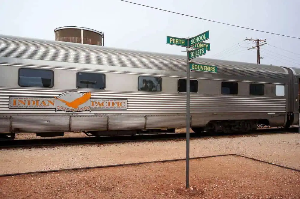 The Indian Pacific train parked at a station with signs pointing to Perth and Sydney. This image relates to the blog post about travel options between Perth and Adelaide, showcasing one of the iconic transport methods across this distance.