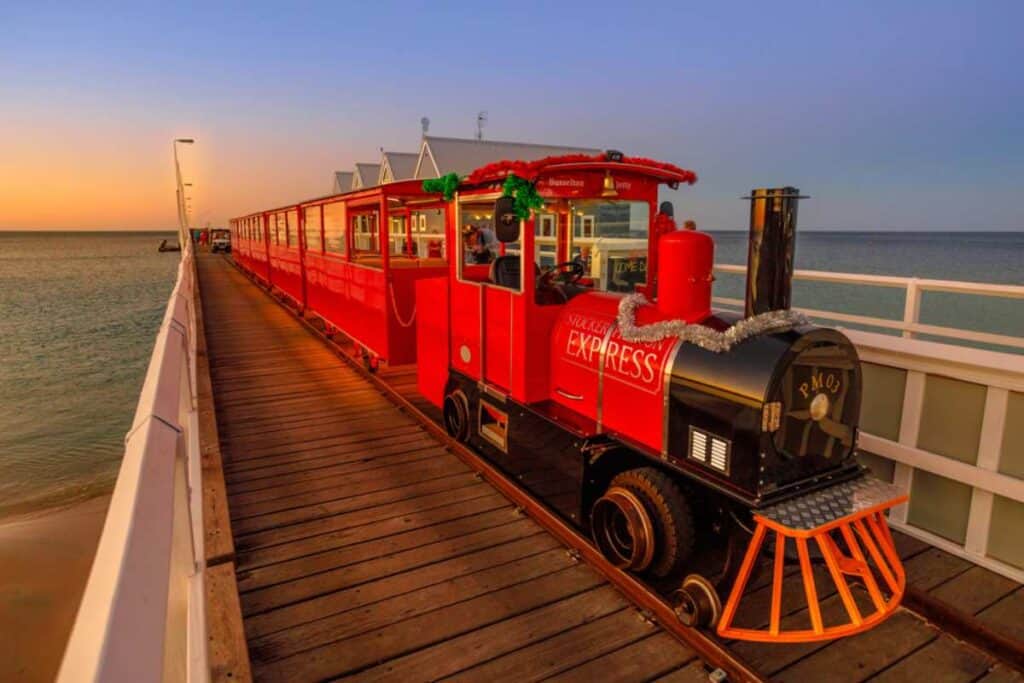 Red 'Stockers Express' jetty train carrying passengers along the Busselton Jetty at sunset. This unique train ride is a popular attraction on the jetty, offering stunning views of the surrounding sea and sky, embodying the charm of Australia’s jetty experiences.