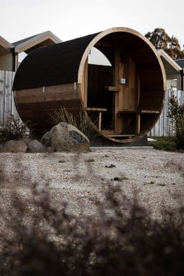 A striking wooden barrel sauna stands on a gravel bed, framed by natural rock landscaping, illustrating the unique accommodation experience offered near Derby, blending luxury with innovative design for a memorable stay.