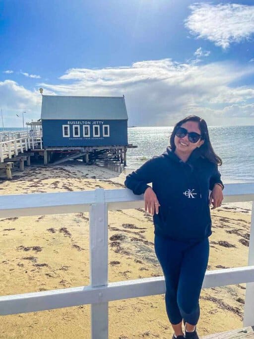 Kate standing on the Busselton Jetty in Australia, smiling and wearing a dark hoodie and sunglasses on a sunny day. The image showcases the longest jetty in Australia, relevant to the post about Busselton Jetty's attractions and experiences.