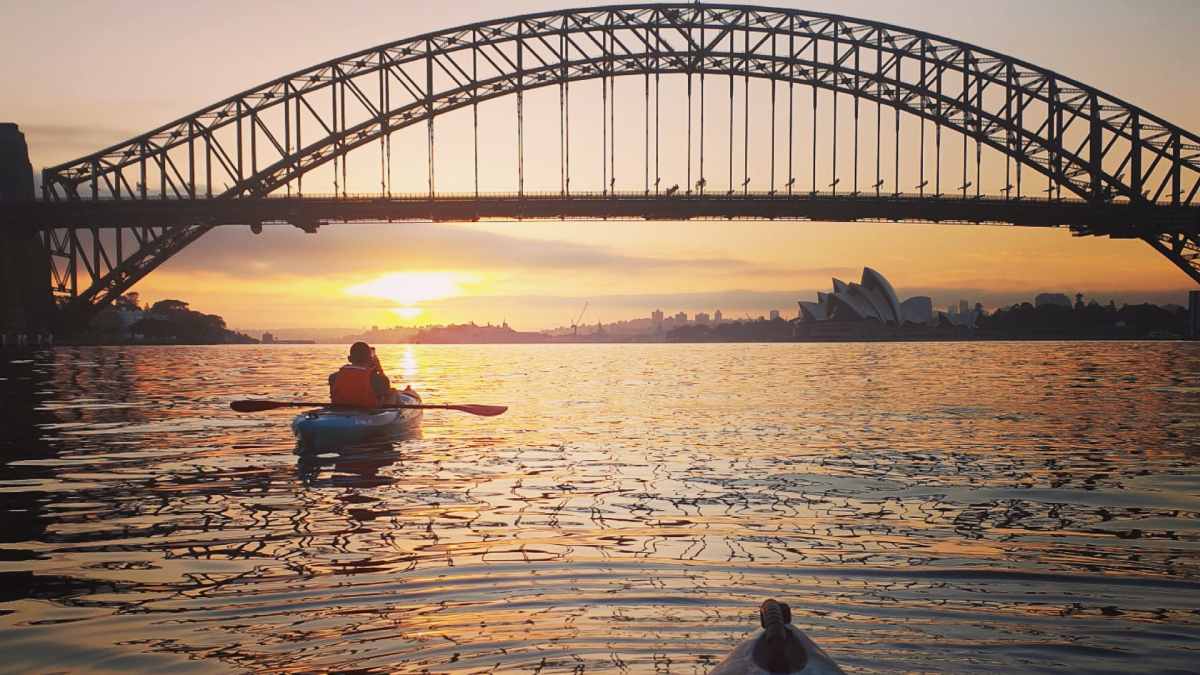 Person kayaking at sunrise on Sydney Harbour with the Sydney Harbour Bridge and Sydney Opera House in the background, highlighting an activity included in many Sydney travel deals.