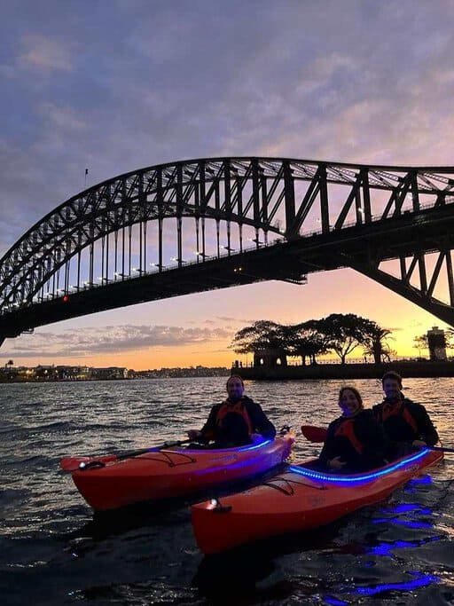 Group kayaking in Sydney under the illuminated Harbour Bridge at dusk, featuring vibrant LED lights on the kayaks.