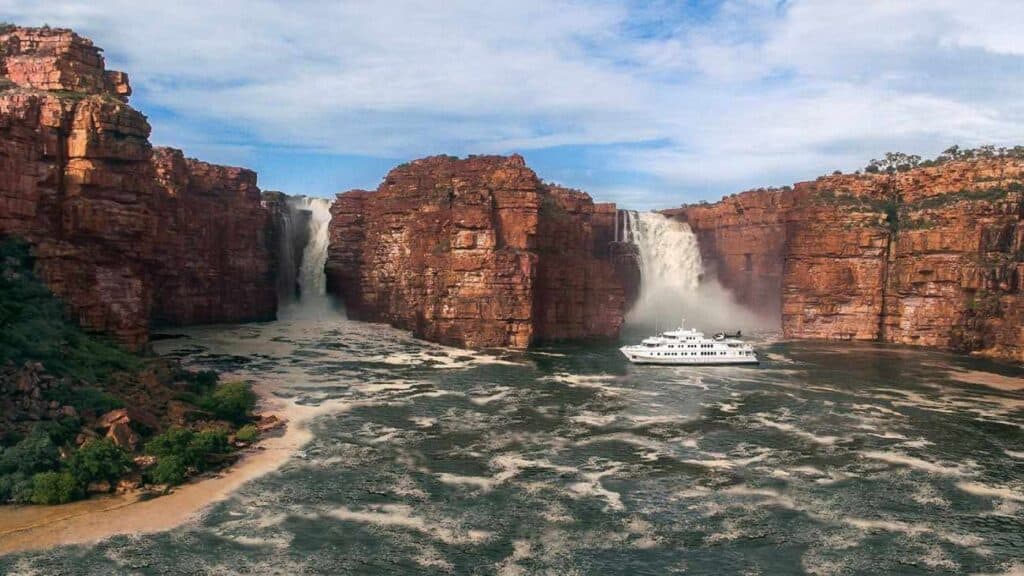 A luxury cruise ship approaching the twin cascades of King George Falls in the Kimberley region of Western Australia, where waterfalls in Australia meet the rugged, red sandstone cliffs, creating a dramatic and unforgettable landscape.