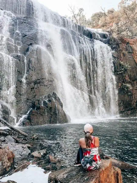 A person sits on a rock, gazing at the stunning Mackenzie Falls in the Grampians, where cascades of water tumble down a rugged cliff into a serene pool, capturing the essence of this popular hiking destination. This is one of the things to do in the Grampians