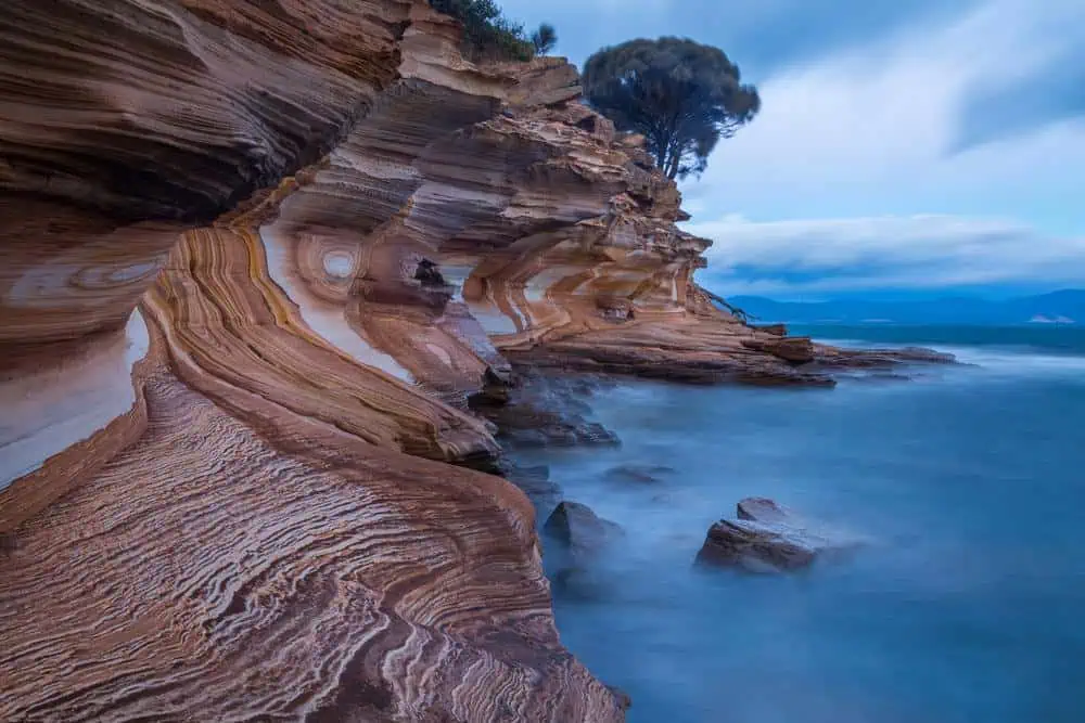 Layered sandstone cliffs on Maria Island, Tasmania, with swirling rock formations meeting the misty shoreline under a cloudy sky. This geological wonder is one of the hidden Tasmania gems, offering a breathtaking coastal landscape for hikers and nature lovers.
