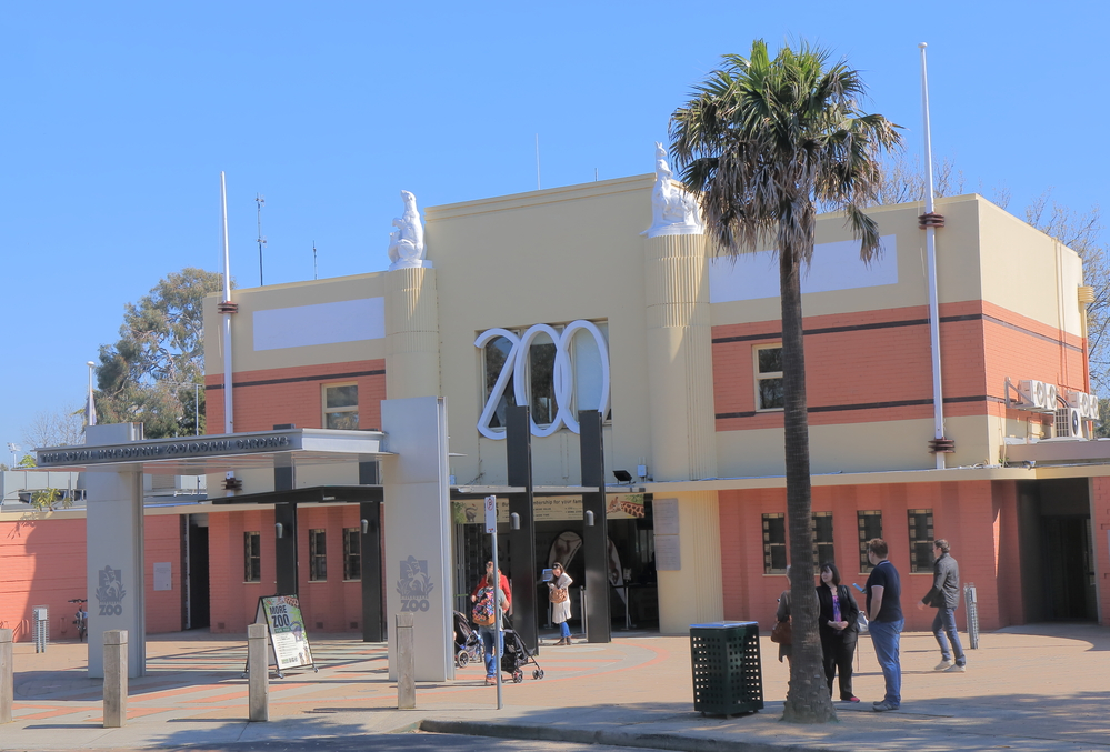A vibrant zoo entrance under a clear blue sky, part of any Melbourne itinerary, features red and beige art deco architecture with large ZOO lettering. People, some with strollers, gather near the entrance. A tall palm tree graces the foreground.