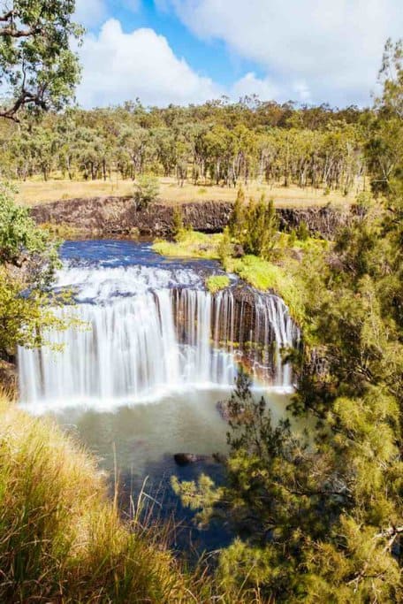 Millstream Falls, with a wide waterfall cascading over a rocky ledge into a serene pool below, surrounded by lush greenery. This stunning sight is one of the best waterfalls to visit in Queensland, making it a must-see destination for nature lovers and adventure seekers.