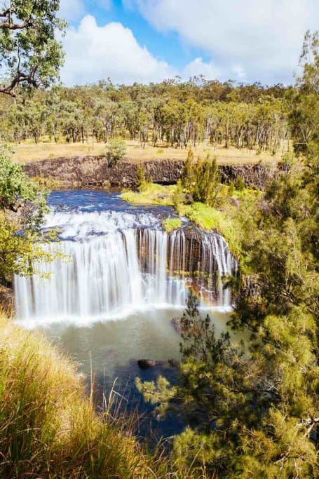Millstream Falls, with a wide waterfall cascading over a rocky ledge into a serene pool below, surrounded by lush greenery. This stunning sight is one of the best waterfalls to visit in Queensland, making it a must-see destination for nature lovers and adventure seekers.