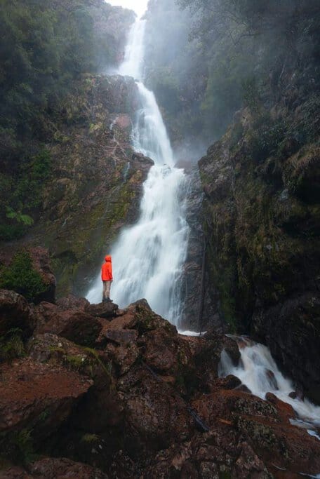 A hiker in a bright orange jacket stands on rocky terrain, gazing up at Montezuma Falls, Tasmania’s tallest waterfall, surrounded by misty rainforest. This stunning natural wonder is one of Tasmania’s best-kept secrets, offering an unforgettable adventure through lush wilderness.