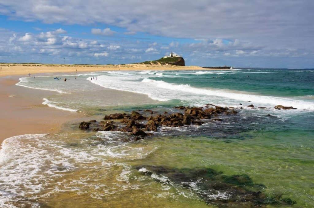 Swimmers and surfers enjoying the waves at Nobbys Beach in Newcastle, with the iconic headland and lighthouse in the distance—one of the coastal highlights on a drive from Sydney to Byron Bay.
