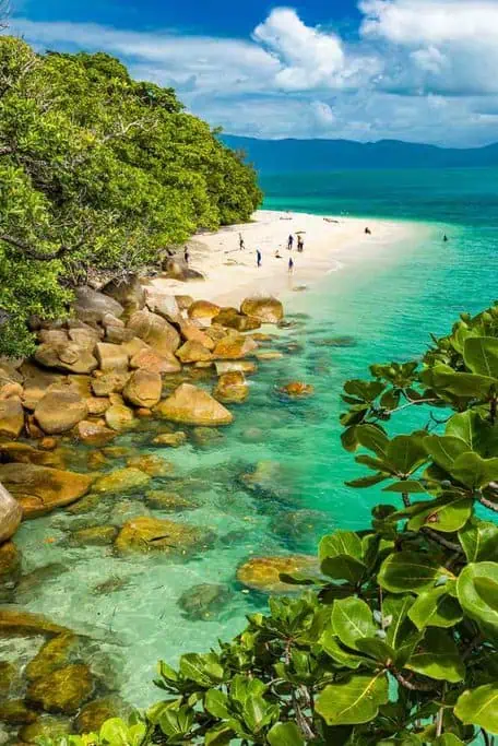 Visitors enjoying the clear turquoise waters and white sandy shore of Nudey Beach, with lush greenery and large boulders, showcasing the natural beauty of secluded beaches in Australia.