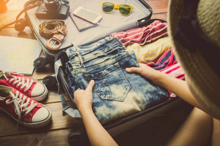 A person packing denim shorts and other clothing into a suitcase, surrounded by travel essentials like shoes, sunglasses, a phone, and a map. The image illustrates the process of preparing for a trip, highlighting smart packing choices for travelling to Australia.