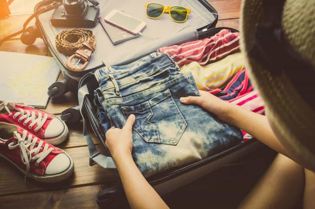 A person packing denim shorts and other clothing into a suitcase, surrounded by travel essentials like shoes, sunglasses, a phone, and a map. The image illustrates the process of preparing for a trip, highlighting smart packing choices for travelling to Australia.