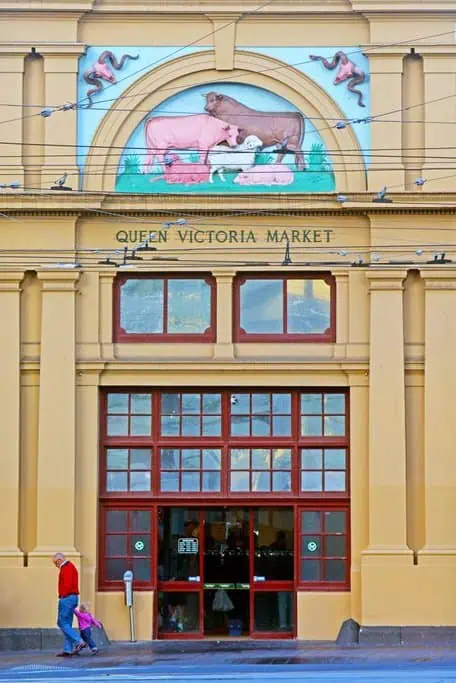 The entrance of Queen Victoria Market, a staple on any Melbourne itinerary, boasts large doors and windows. Above, a mural depicting cows and sheep captivates visitors. A person in a red top and hat strolls by as pigeons perch on the ledge.