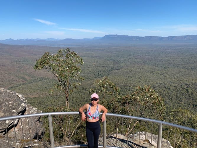 I stand at Reed Lookout in the Grampians, smiling at the camera with The Balconies' vast, forested landscape stretching into the distance under a clear sky.