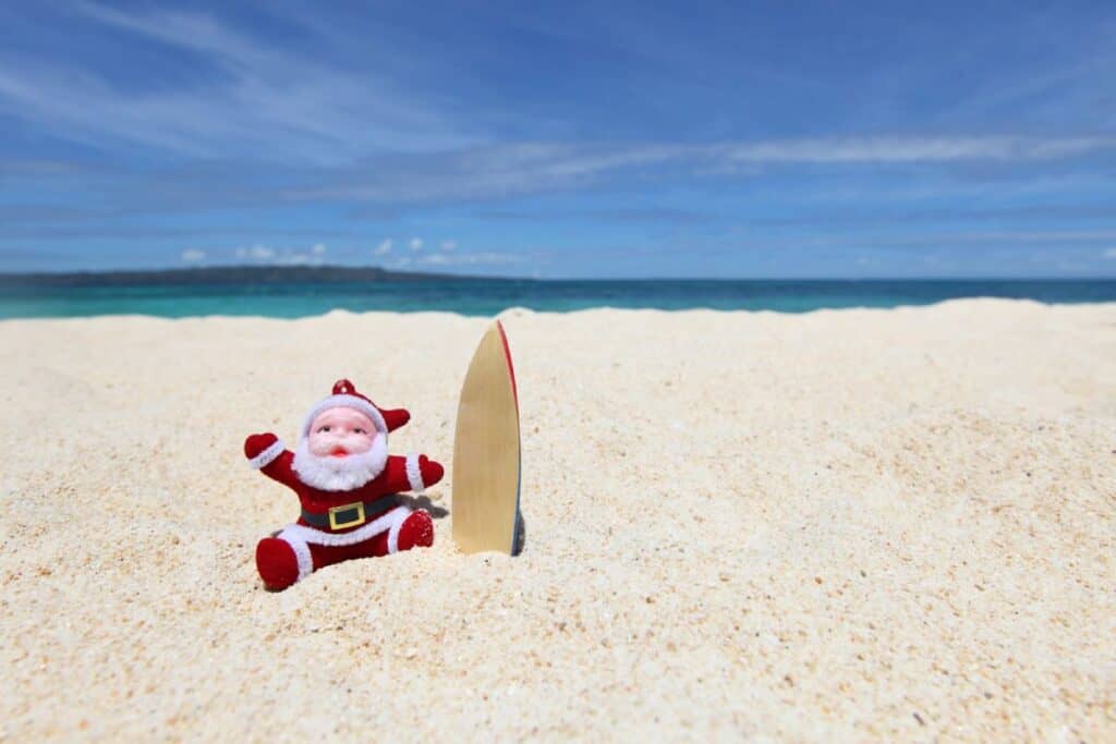 A Santa Claus figurine sitting on a tropical beach with a small surfboard, representing a quirky take on Christmas in Australia. One of the cool facts about Christmas in Australia is the combination of traditional holiday symbols with summer beach culture, where Santa might be seen enjoying the surf instead of snow.