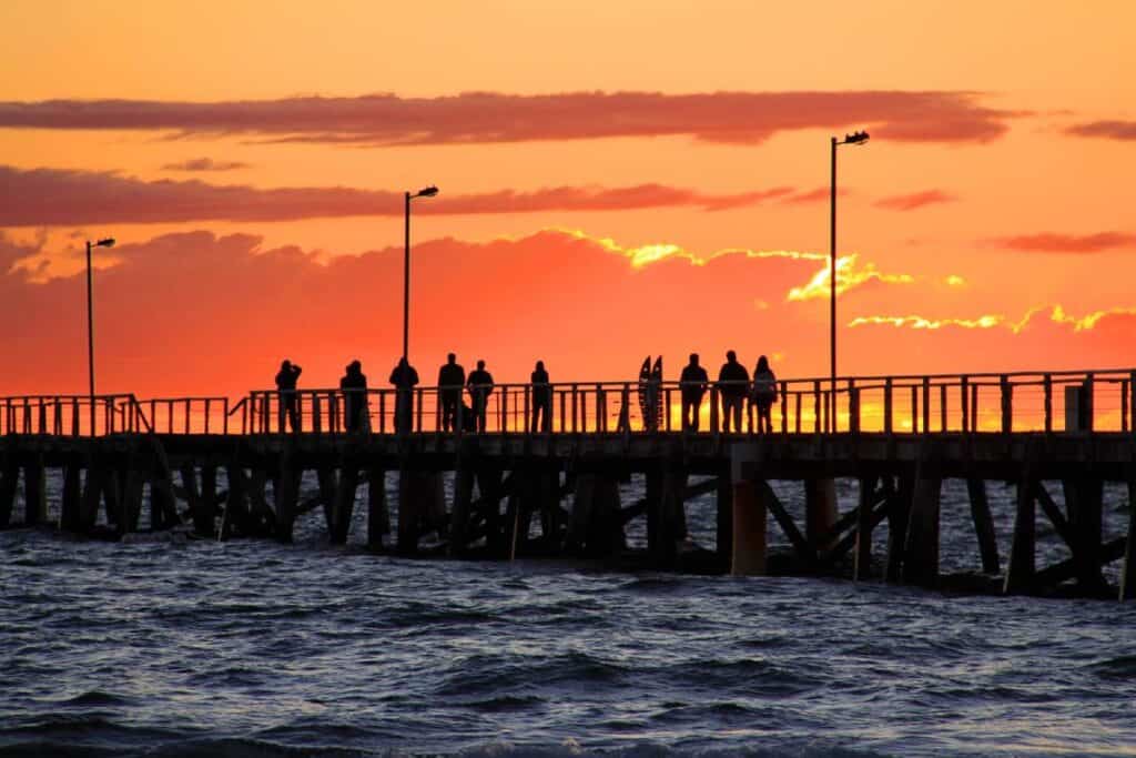 People standing on Semaphore Jetty in Adelaide at sunset, capturing the serene end of a day and connecting to the distance from Adelaide to Perth.