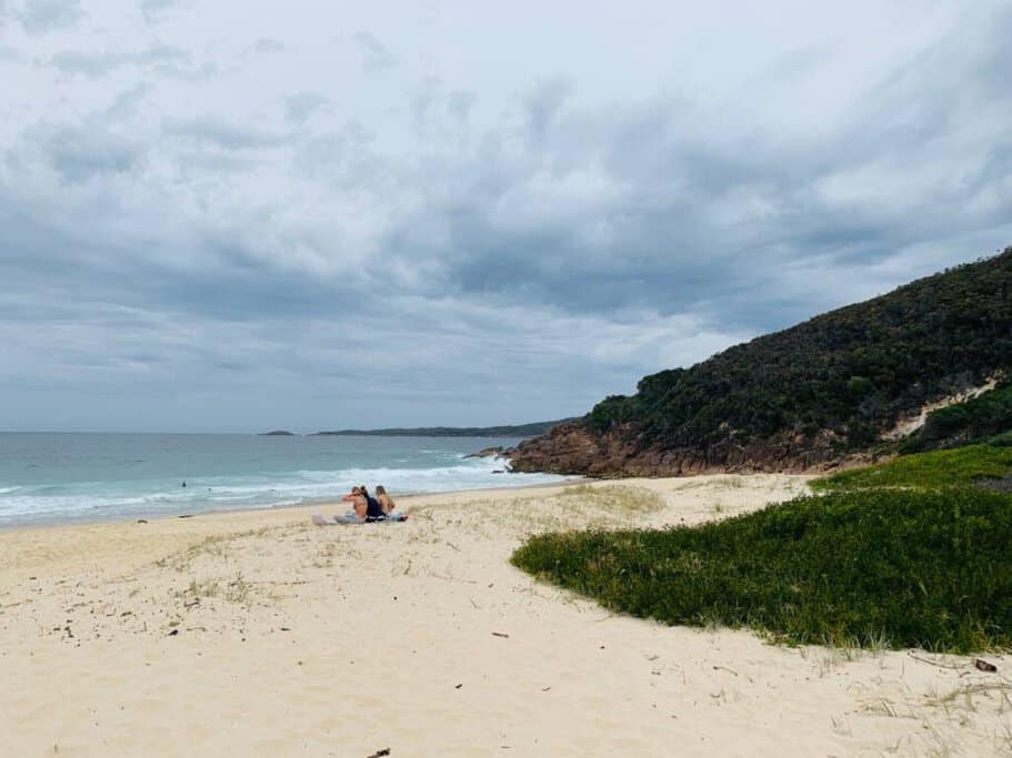 Visitors relaxing on the sandy shore of Shoal Bay, one of the famous beaches in Port Stephens, with lush greenery and rocky headlands in the background, capturing the beach's natural beauty and serene atmosphere.
