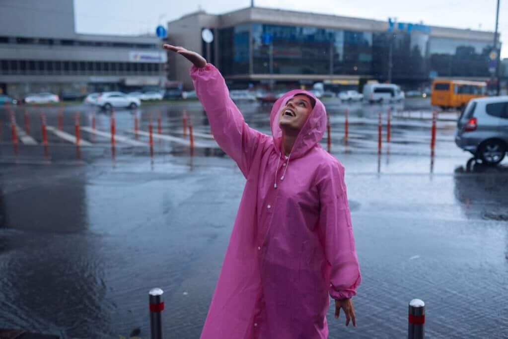 A woman wearing a pink raincoat smiles and enjoys the rain in an urban setting. This image relates to the idea of packing for unpredictable weather in Australia, highlighting the importance of being prepared with appropriate clothing like rain gear when travelling