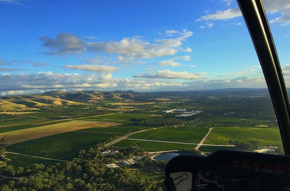 A panoramic view from a helicopter cockpit showing the sprawling vineyards and undulating hills of the Barossa Valley at sunset. The rich tapestry of greens and browns below, interspersed with roads and farm buildings, illustrates the scenic flights over this renowned wine region, offering a breathtaking perspective that encapsulates the essence of helicopter tours across the Barossa Valley.